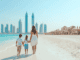 Dubai mit Kindern. EIne Mutter geht mit ihren Kindern über den weissen Sandstrand. Im Hintergrund ist die Skyline von Dubai zu sehen. rechts ist ein Beach Club