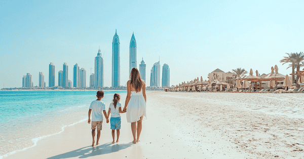 Dubai mit Kindern. EIne Mutter geht mit ihren Kindern über den weissen Sandstrand. Im Hintergrund ist die Skyline von Dubai zu sehen. rechts ist ein Beach Club